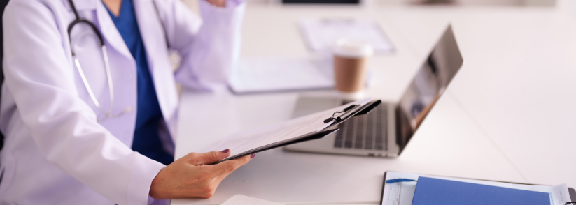 Healthcare professional holding clipboard in front of laptop, notebook, and coffee at desk